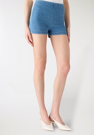 Kobieta w niebieskich krótkich spodenkach z wysokim stanem w białe kropki i białych niskich butach na obcasie na jasnym, prostym tle.