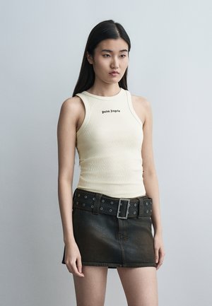 Mladá žena s dlhými čiernymi vlasmi má na sebe krémový tielko s potlačou a čiernu džínovú minisukňu so širokým opaskom.