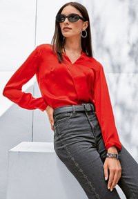 Rode, langemouwen, zijden blouse met een V-hals, gecombineerd met hoge taille, donkere grijze jeans met een verticale lijn van versieringen. Zwarte zonnebril.