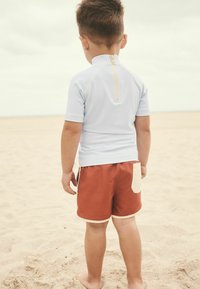 Krátký rukáv světle modrý rash guard se zipem na zádech, spárovaný s hnědými plavkami s krémovou kapsou a bílým lemem. Písečné pozadí.