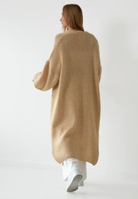 Beżowy, oversize, dzianinowy kardigan o teksturowym wykończeniu, luźnymi rękawami i asymetrycznym dołem. Noszony na jasne spodnie i trampki.