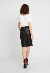 Jupe crayon en cuir noir avec une texture lisse, longueur genoux, comportant une couture au centre de l'arrière et une fermeture éclair dissimulée. Associée à un t-shirt blanc.