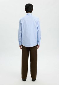 Man die met zijn rug naar ons toe staat, gekleed in een lichtblauw overhemd met knopen en een donkerbruine broek met wijde pijpen, op een effen witte achtergrond.