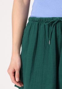 Gonna in tessuto verde, con texture, dotata di una cintura elastica e lacci. Il tessuto sembra morbido e traspirante, scivolando liberamente attorno alla vita.