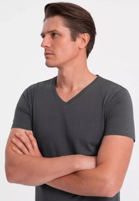 Šedé tričko s krátkým rukávem a výstřihem do V vyrobené z hladké bavlny. Model stojí se zkříženýma rukama, což zvýrazňuje vykrojení a střih trička.