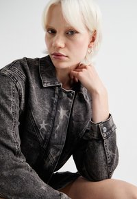 Juodas džinsinis švarkas su nusidėvėjusiu paviršiumi ir žvaigždžių raštu, su apykakle, mygtukų užsegimu ir rankogalių detalėmis. Lygus paviršius, kasdieniškas dizainas.