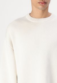Pulover alb din tricot, cu guler rotund ribflat, având un model texturat și o croială cu umeri căzuți pentru o ajustare relaxată.