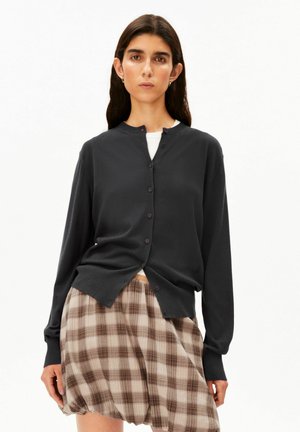 Frau, die eine schwarze, geknöpfte Strickjacke über einem weißen Top und einem beige-braunen karierten Rock trägt, steht vor einem schlichten weißen Hintergrund.
