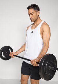 Débardeur de sport sans manches blanc avec un logo noir, associé à un short noir, tenant une barre noire avec des poids ronds.