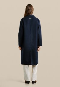 Marineblauwe jas met een kraag, lange mouwen en een rechte snit. Heeft een centrale naad en een label aan de achterkant. Draagt over witte broek.