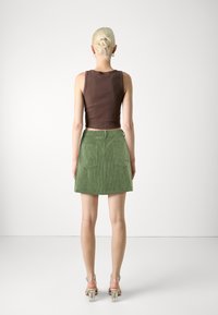 Débardeur marron court associé à une jupe en velours côtelé vert avec deux poches arrière, vu de dos. Sandales à talons blocs transparents.