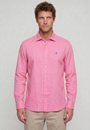 Hombre con camisa rosa de manga larga con pequeño logo azul en el pecho, combinado con pantalones beige, fondo neutro.