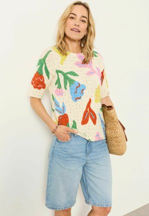 Frau trägt einen bunten kurzärmeligen Pullover mit floralem Muster und helle Denim-Shorts bis zum Knie, hält eine geflochtene Strohtasche vor weißem Hintergrund.