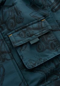 Mörk teal-tyg med ett livligt mönster av byggfordon. Har en ficka med lock och en texturerad knappstängning samt orange sömmar.