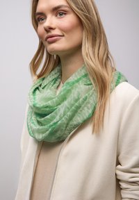 Écharpe à motifs vert clair drapée autour du cou, en tissu transparent. Portée avec une veste crème texturée et un haut beige.