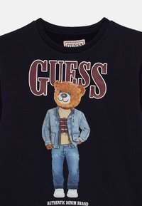 Μαύρο βαμβακερό t-shirt με σχέδιο αρκούδας που φοράει τζιν μπουφάν, μπλε τζιν και άσπρα αθλητικά παπούτσια, με την επιγραφή "GUESS" επάνω.