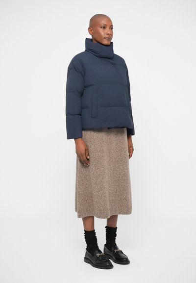 Tumes sinine puff-jakk kõrge kaelusega, ees oleva tasku ja lühikese disainiga, kantud beeži tekstuuriga kudumisseeliku ja mustade loafersitega, millel on kuldsed aktsendid.