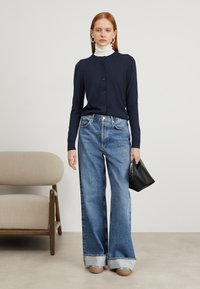 Mornarska jopica na gumb, v kombinaciji s svetlo modrimi širokimi hlačami s manšetami. Model drži črno torbico in nosi belo polo majico.