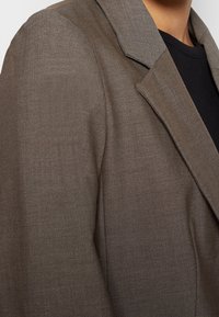 Ruskea bleiseri, jossa on teksturoitu kangas, terävä kaulus ja muotonsa mukautettu istuvuus. Suunnittelu sisältää siistin siluetin, jossa on minimaalista yksityiskohtaisuutta.