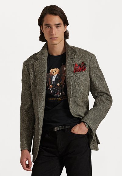 Sako s houndstooth vzorom v hnedej a béžovej farbe s náprsním vreckom v červenej kocke, nosené over grafickým čiernym tričkom a spárované s tmavými džínsami.