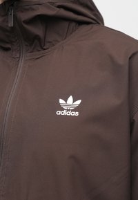 Καφέ μπουφάν με φερμουάρ μπροστά, διακοσμημένο με το λευκό λογότυπο τριφυλλιού Adidas στο στήθος, φαίνεται με κουκούλα προσαρτημένη.