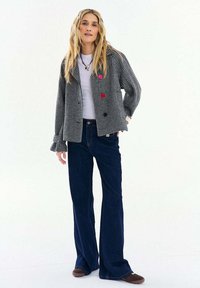 Žena s dlhými blond vlasmi, ktorá má na sebe čierno-bielu károvanú bundu s farebnými gombíkmi, biele tričko, tmavomodré zvonové džínsy a hnedé topánky.