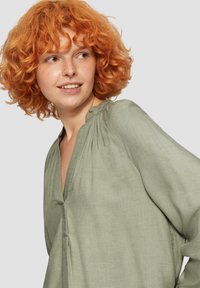 Hellgrüne Bluse mit langen Ärmeln und V-Ausschnitt, die an den Schultern geraffte Akzente und eine weiche, fließende Stofftextur aufweist.