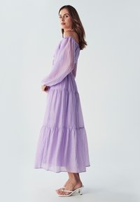 Laventelin värinen kerroksellinen maksimekko, jossa on läpinäkyvät pitkät hihat ja olkaimeton kaulus, sekä pehmeä rakenne ja ilmava muotoilu. Yhdistettynä valkoisiin sandaaleihin.