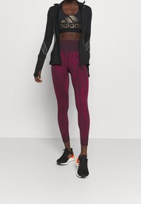 Black zip-up jacket, gold logo sports bra, and maroon patterned leggings paired with black athletic shoes featuring orange accents.