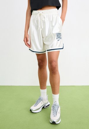 Person iført hvide Nike basketballshorts med navy trim og hvide Nike sneakers med grå accenter på grøn gulv.