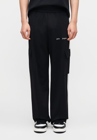 Μαύροι cargo παντελόνια με ελαστική μέση, πλαϊνές τσέπες, λογότυπο μπροστά και αντίθετες πλαϊνές πιέτες. Φορεμένα με μαύρα και λευκά αθλητικά παπούτσια.