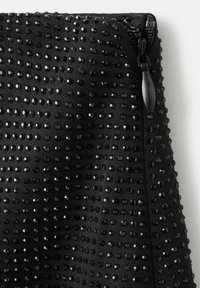 Sorte lynlåsesektion til beklædning med et struktureret stof, hvor små, blanke pailletter er arrangeret i et regelmæssigt mønster.