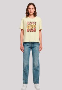 Light yellow cotton t-shirt featuring bold, colorful "BEST MOM EVER" text and floral accents, paired with blue jeans and white sneakers.