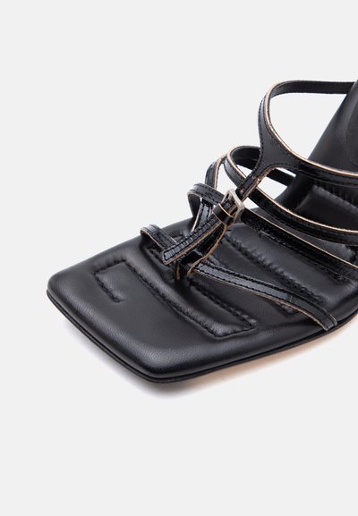 Μαύρο δερμάτινο σανδάλι με τετράγωνη μύτη, λεπτά διασταυρούμενα λουράκια, μικρό αγκράφα και επενδυμένο πέλμα σε λευκό φόντο.