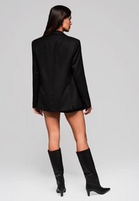 Černý střihový blazer se strukturovaným designem, zapínáním na jedno knoflík a dlouhými rukávy. V kombinaci s černými vysokými kozačkami na podpatku.