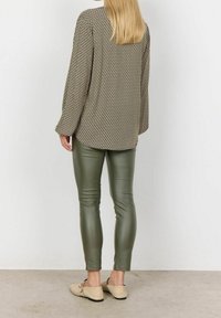 Langarmige, beige gemusterte Bluse mit lockerem Schnitt, kombiniert mit enganliegenden olivgrünen Kunstlederhosen und beigen Slippern.