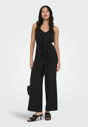 Žena oblečená v čiernom overal bez rukávov s širokými nohavicami, so sťahovacou páskou v páse, nesie malú čiernu kabelku a má na nohách čierne sandále na podpätku.