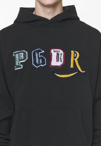 Sort hættetrøje med flerfarvede broderede bogstaver og mønstre, inklusive en banangrafik, med en forlommekangurulomme.