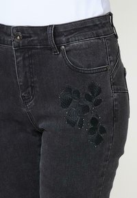 Juodos džinsinės kelnės su gėlių siuvinėjimu kairėje šlaunies pusėje, papuoštos smulkiais akmenukais. Klasikinis penkių kišenių stilius.