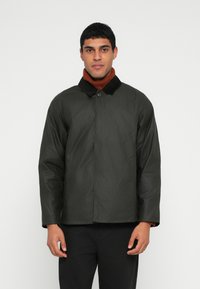 Σκούρο πράσινο αδιάβροχο μπουφάν με μαύρο γιακά, κλείσιμο με κουμπιά και πλαϊνές τσέπες, φορεμένο πάνω από ένα σκουρόχρωμο ψηλόμεσο πουλόβερ καφέ χρώματος.