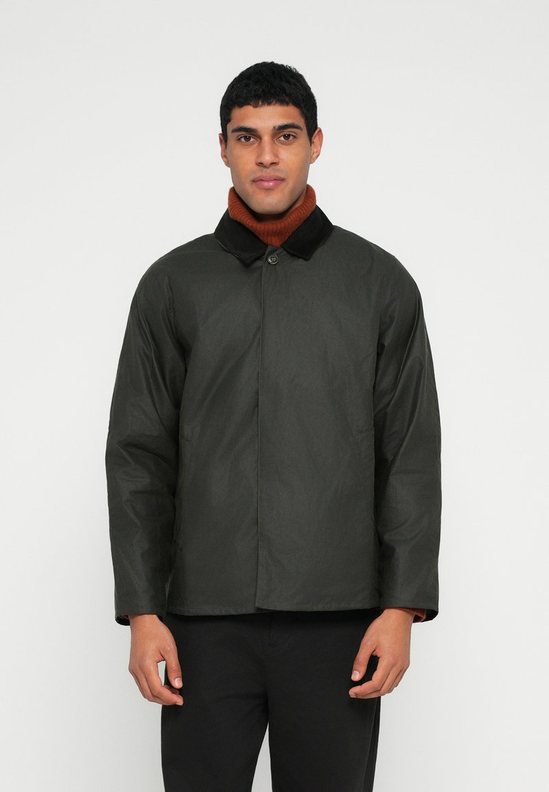 Σκούρο πράσινο αδιάβροχο μπουφάν με μαύρο γιακά, κλείσιμο με κουμπιά και πλαϊνές τσέπες, φορεμένο πάνω από ένα σκουρόχρωμο ψηλόμεσο πουλόβερ καφέ χρώματος.