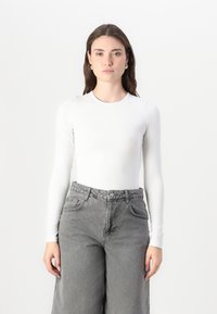 Bílé body s dlouhým rukávem vyrobené z hladké látky, zkombinované s šedými širokými džíny s vysokým pasem, které mají přední kapsy a zapínání na knoflík.