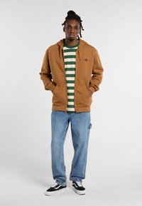 Brown zip-up hoodie with front pockets over a green and white striped t-shirt, paired with blue jeans and black sneakers.