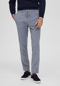 Pantaloni grigi su misura con un delicato motivo a quadri, vita con cordoncino e tasche laterali, abbinati a sneakers scure.