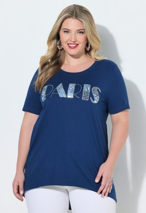 Smilende kvinde med langt blondt hår iført en blå T-shirt med pailletter og teksten "PARIS" samt hvide bukser, stående mod en ensfarvet lys baggrund.