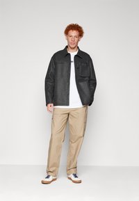 Veste texturée gris foncé avec une poche avant, T-shirt blanc en dessous, pantalon cargo beige clair, et chaussures blanches avec des lacets bleu marine.