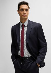 Un costume bleu marine foncé au design ajusté, associé à une chemise blanche à fleurs et une cravate bordeaux. Il présente un tissu texturé et une coupe sur mesure.