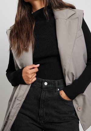 Kvinde iført sort ribbet langærmet top, ærmeløs beige vest og sorte højttaljede jeans med hånden i lommen og let manicure.