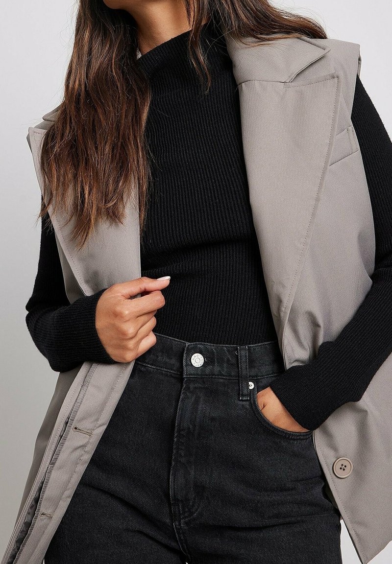 Femme portant un haut noir côtelé à manches longues, une veste beige sans manches et un jean noir taille haute, main dans la poche et manucure légère.