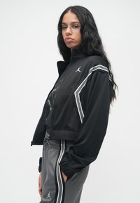 Jauna sieviete ar gariem, cirtainiem melniem matiem, kas nēsā brilles, melnu zip-up jaku un pelēkas sporta bikses ar baltām svītrām, stāv sānis.